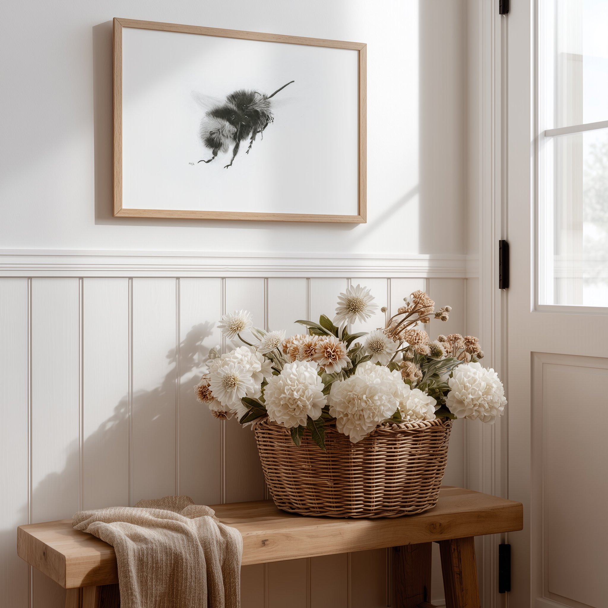 Charcoal bumblebee art print in a light wood frame above white wainscoting, styled with a wicker basket of dried florals—cottagecore pollinator wall art for an entryway.
