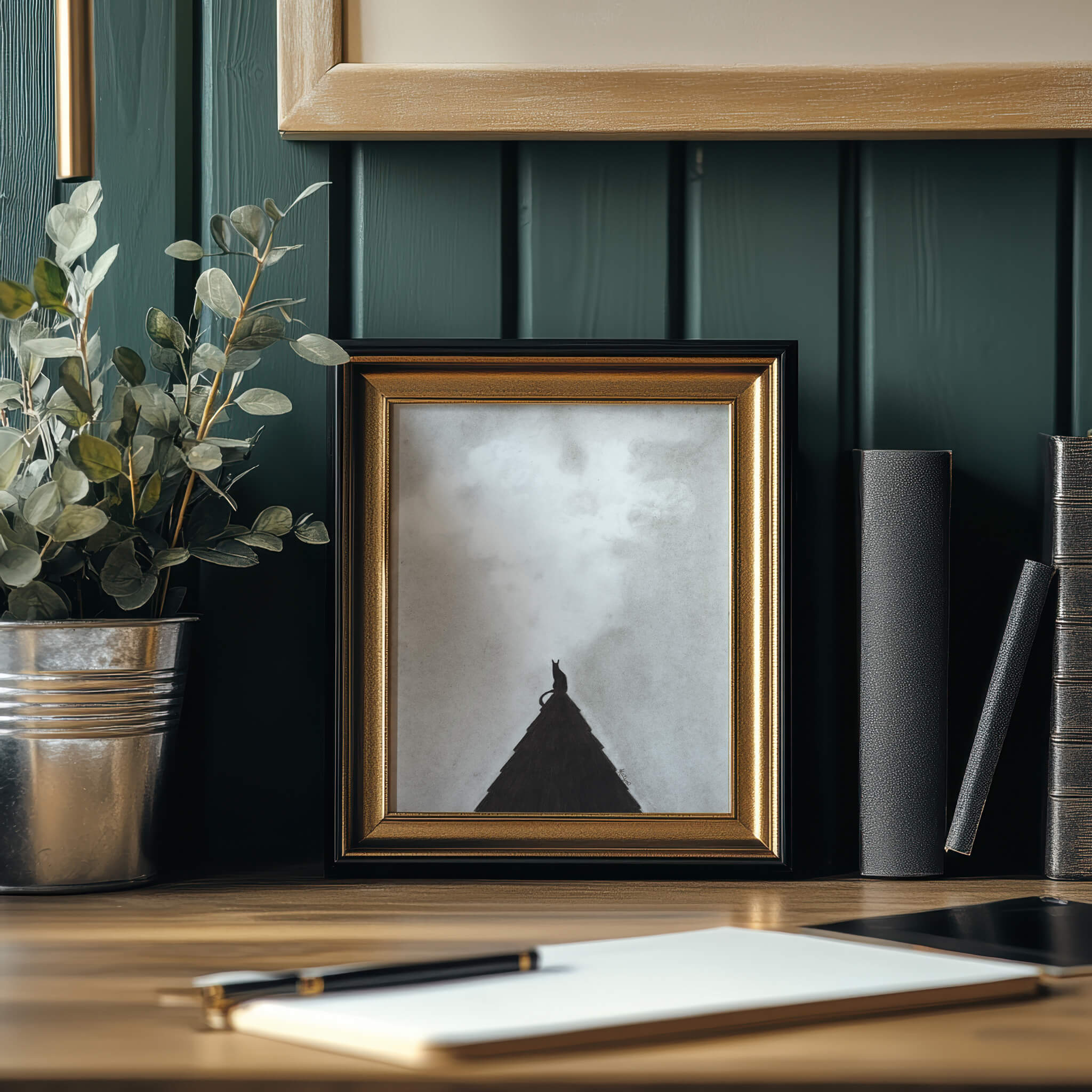 Charcoal black cat art print of a cat perched on the peak of a black roof with a moody sky above, framed in gold and black, leaning against a dark paneled wall styled with books, a plant, a notepad, and a pen.