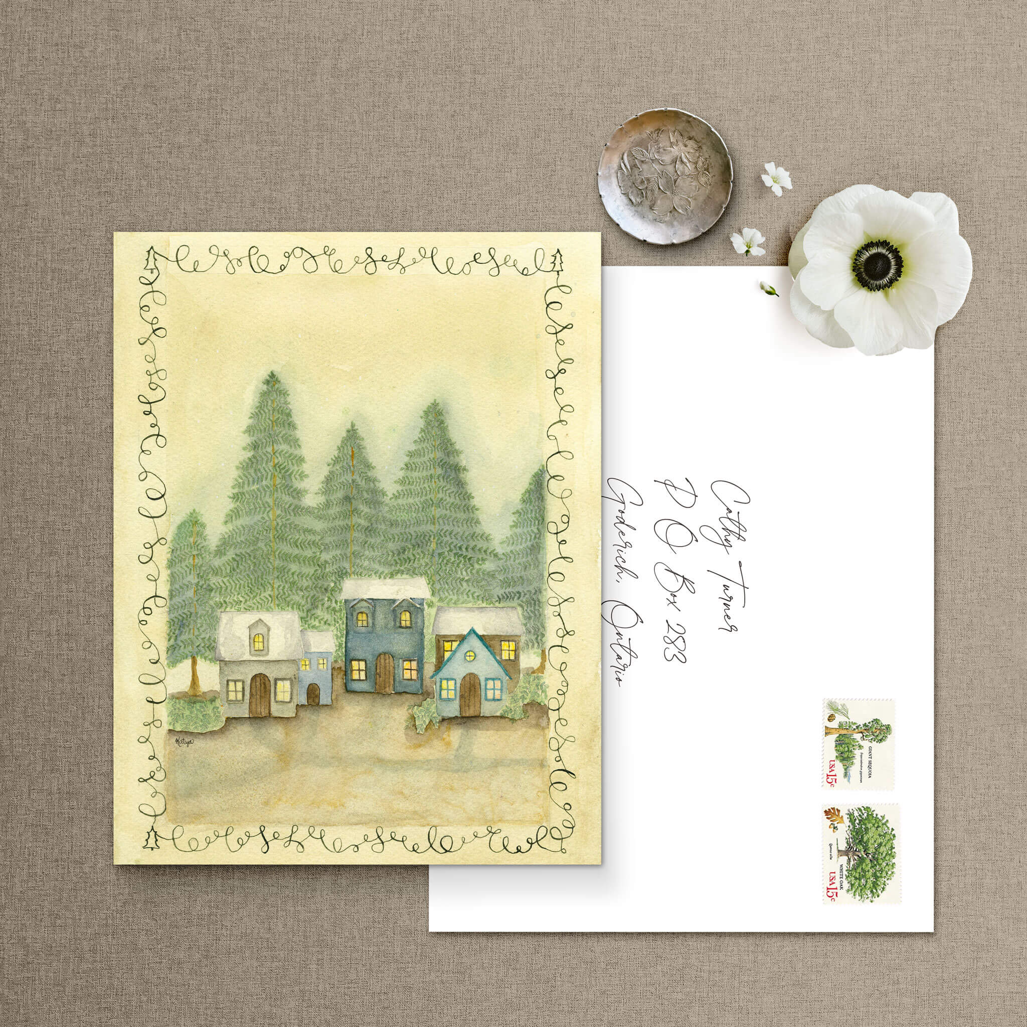 Nestled Among the Trees winter village notecard beside a hand-addressed envelope with vintage tree stamps and a white flowers.