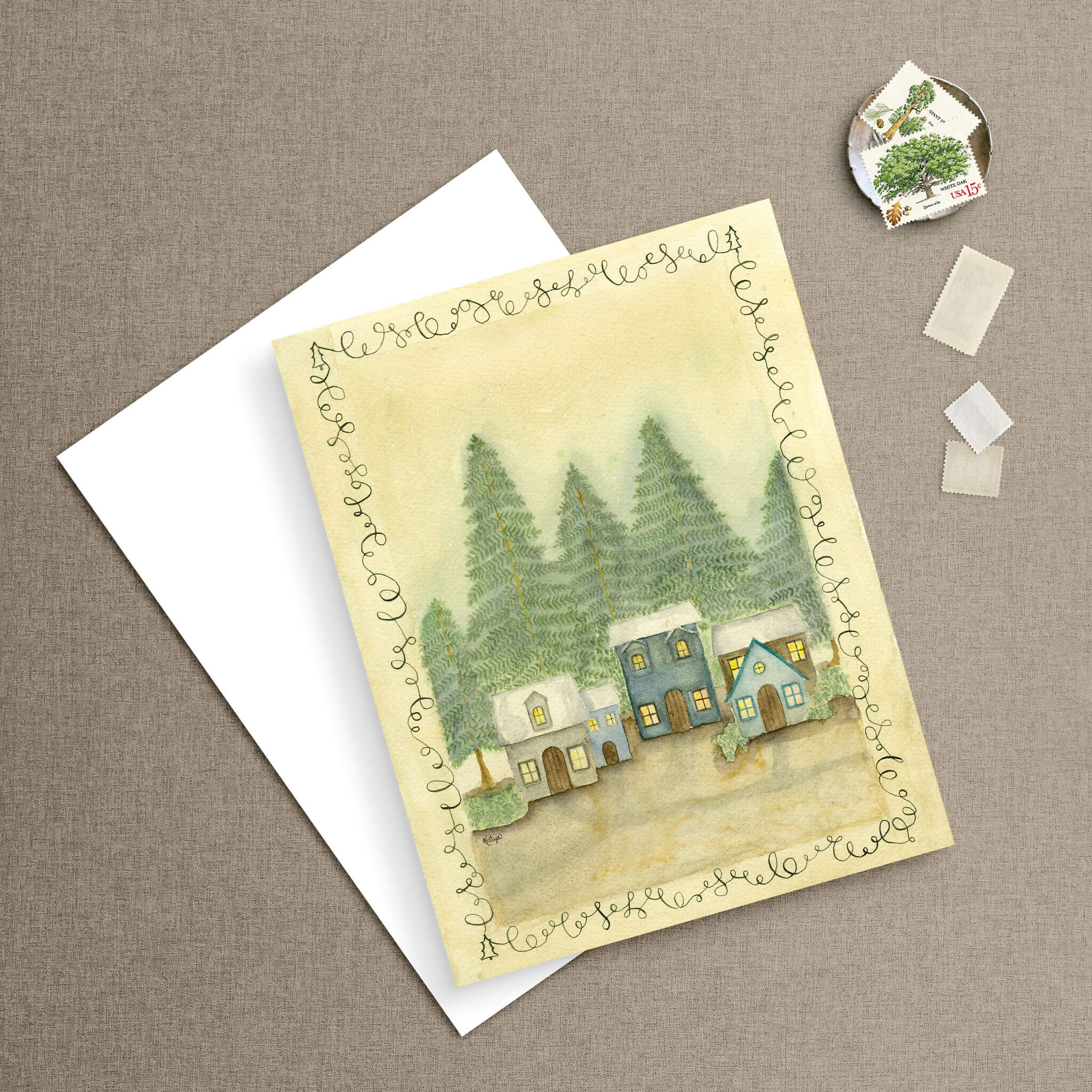 Nestled Among the Trees winter village notecard styled beside a white envelope with vintage tree postage stamps on a linen background