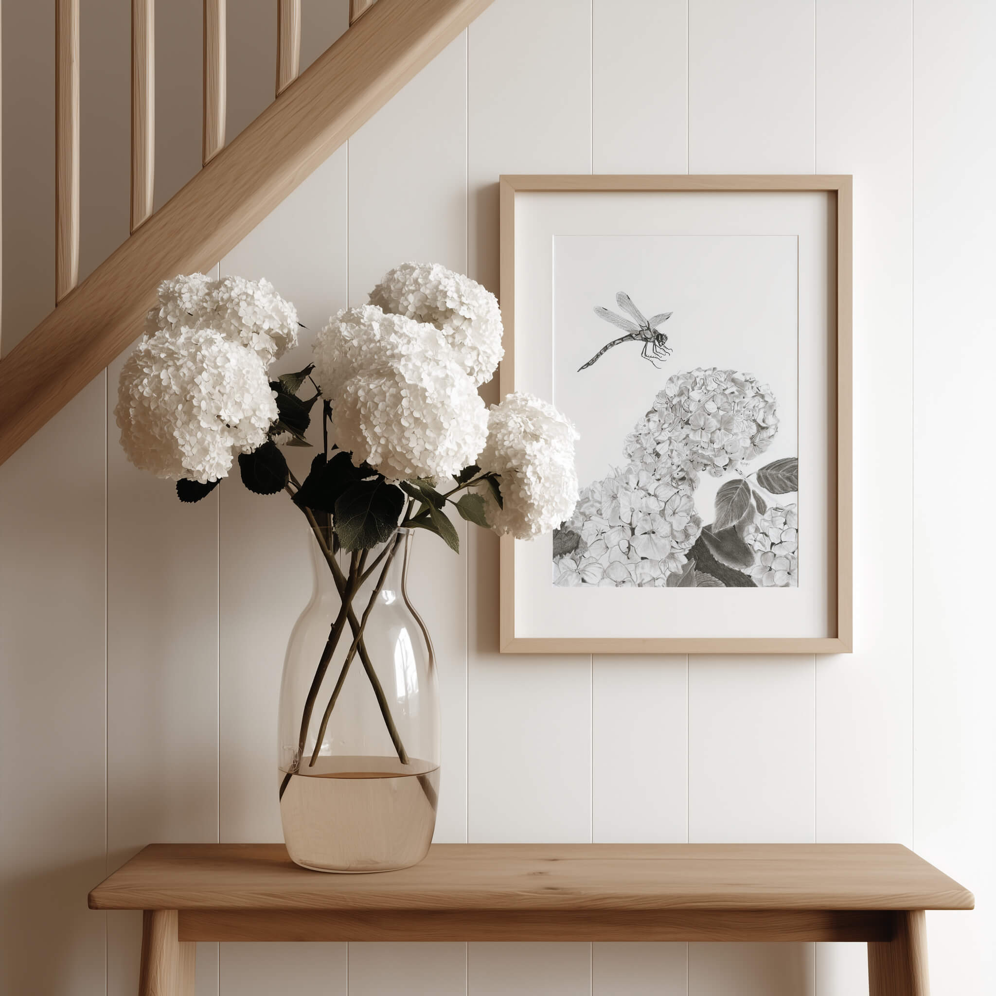 Touchdown dragonfly art print in an oak frame on an entryway console beside a vase of white hydrangeas under a staircase