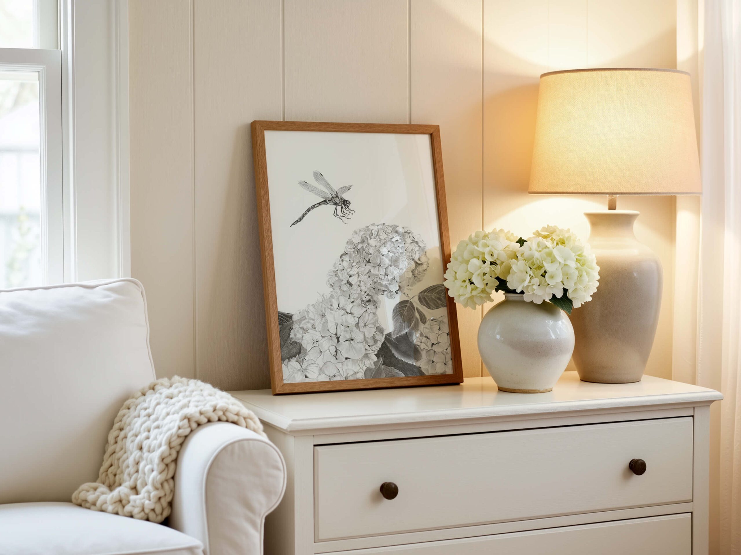 Touchdown dragonfly art print in an oak frame on a cream dresser beside a vase of white hydrangeas