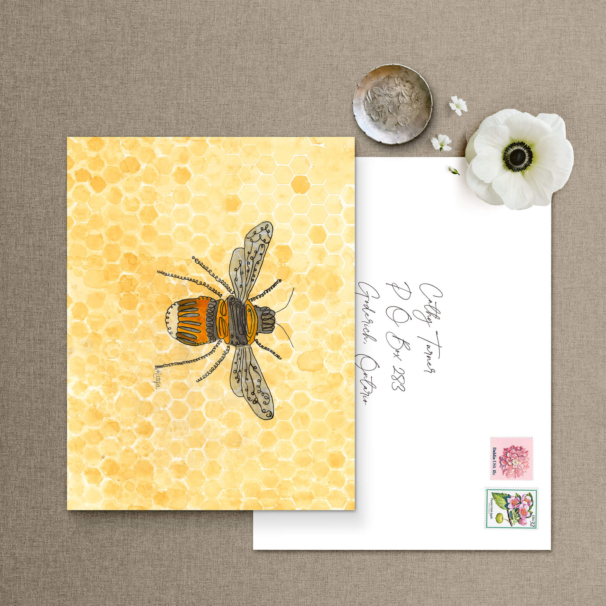 A bee art notecard beside a hand-addressed envelope with vintage floral stamps and a white flower