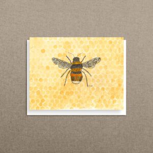 A bee art notecard featuring a watercolor bee on a golden honeycomb background, propped against warm linen.