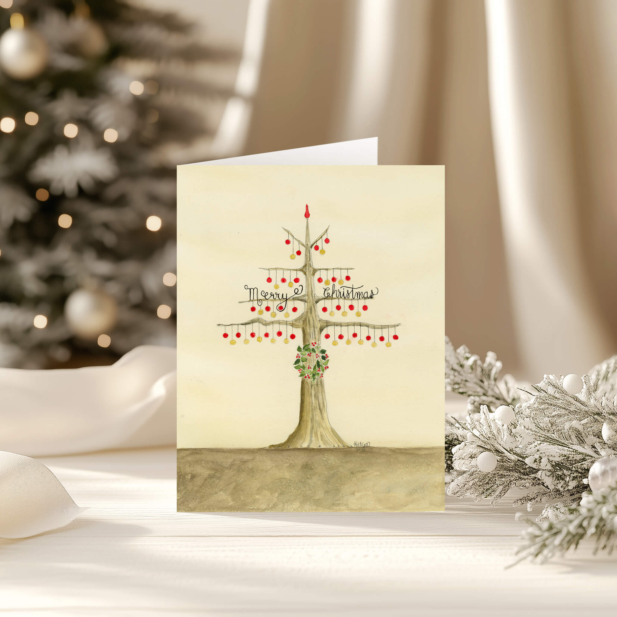 Merry Christmas card with a watercolor tree of red and gold ornaments, standing on a white table beside frosted pine and a glowing tree.