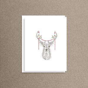 Christmas deer card featuring a deer drawn in swirling black ink loops, its antlers hung with red, green and gold baubles and a red garland, shown with a white envelope on taupe linen