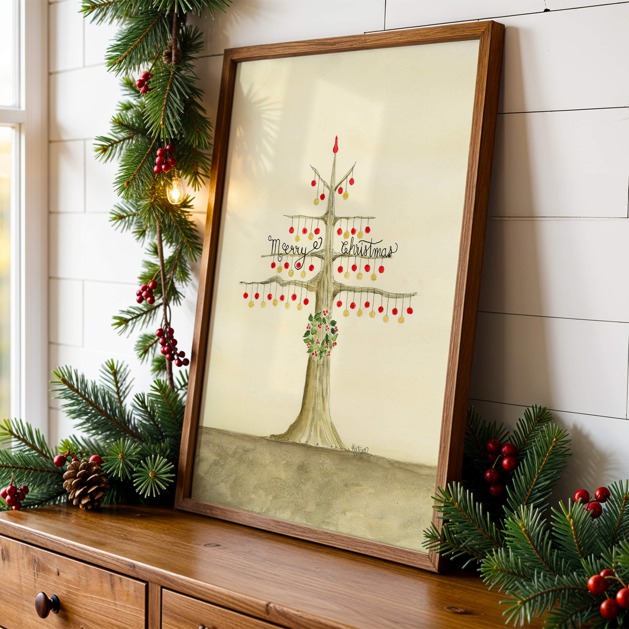 Original Christmas painting of a watercolor tree with red and gold ornaments, framed and leaning on a wooden console among pine garland and red berries