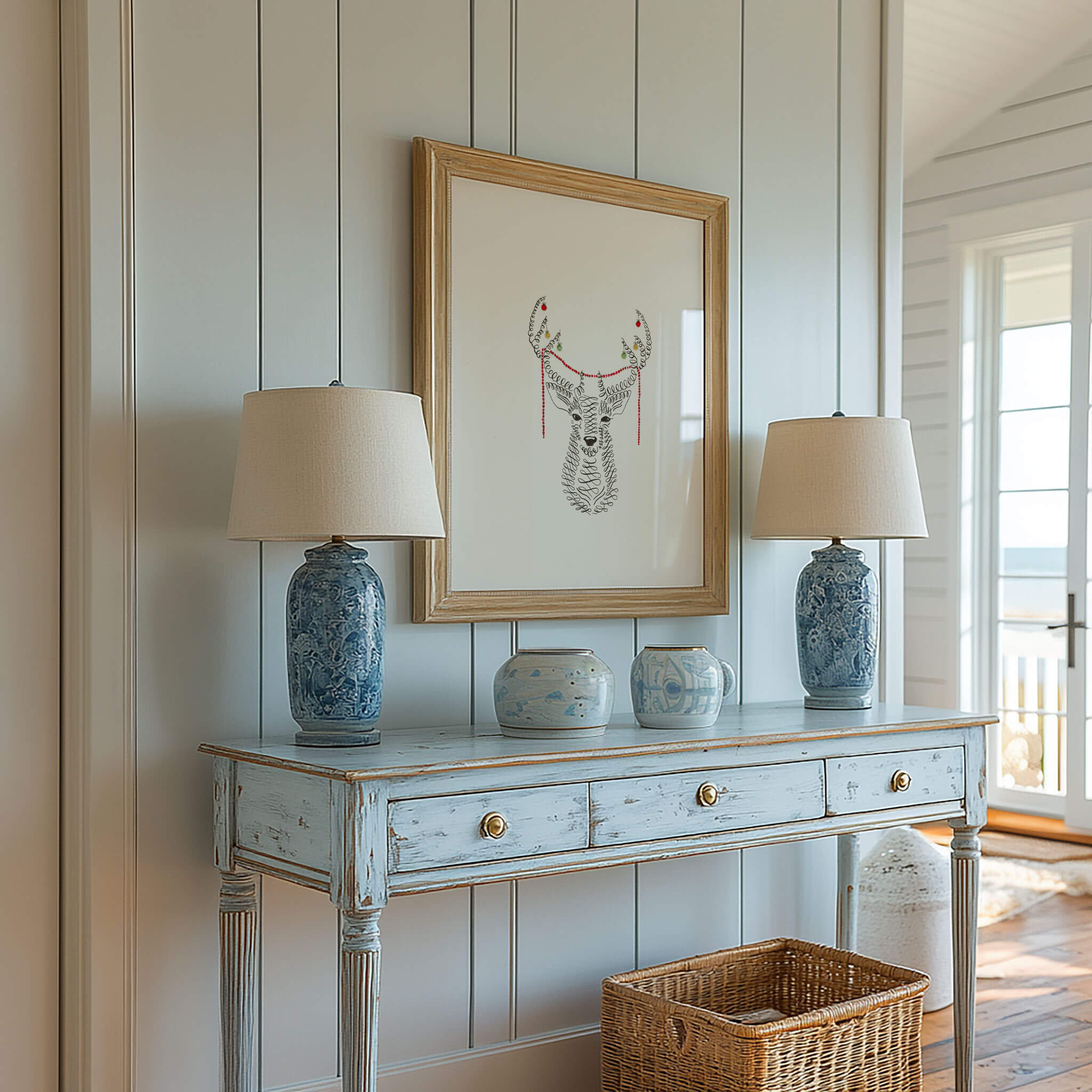 Original pen and ink deer drawing in a weathered wood frame above a distressed blue console with blue and white lamps and a wicker basket, near a sunny ocean-view door.