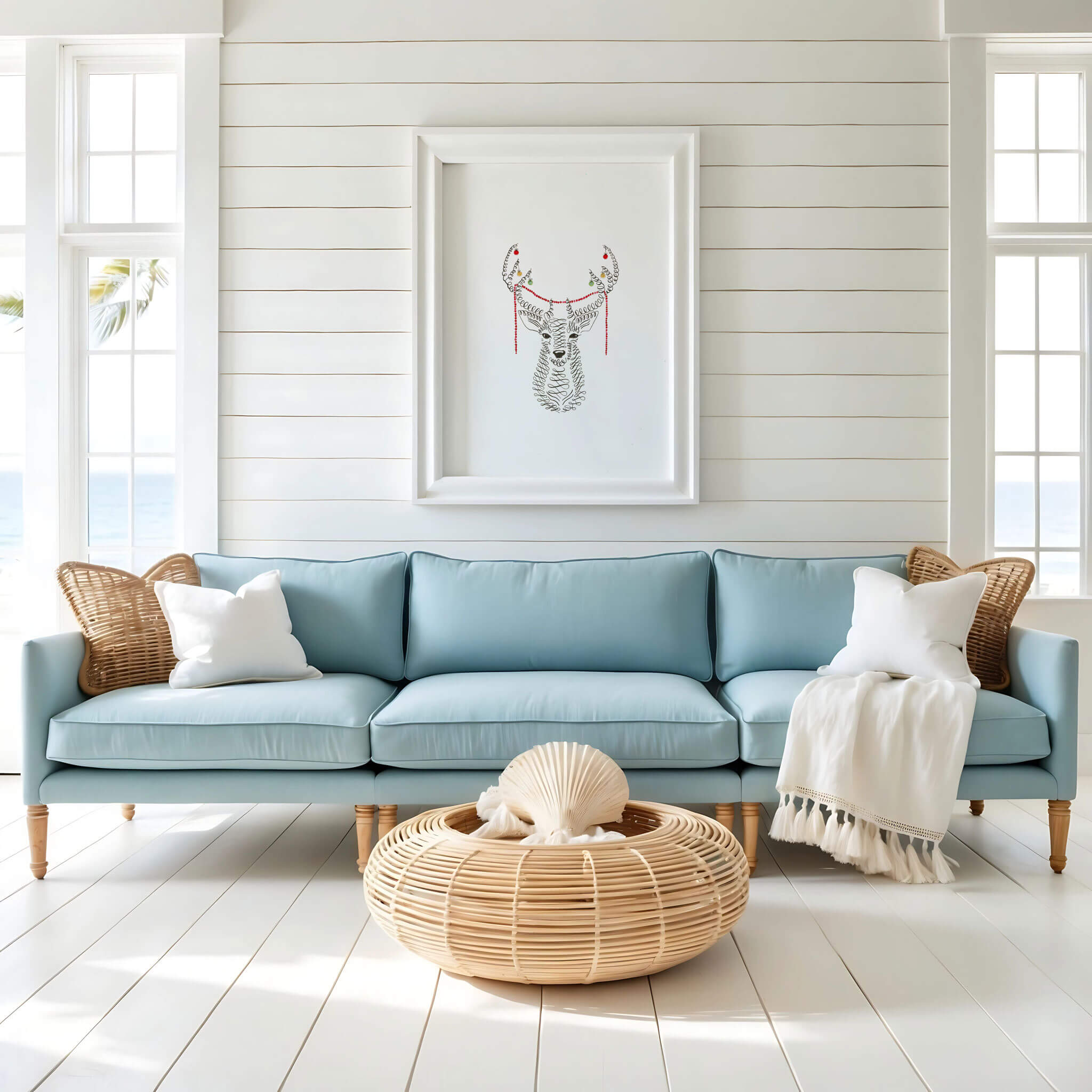 Original calligraphy Christmas deer drawing in a white frame on white shiplap above a pale blue sofa, with rattan accents and ocean views.