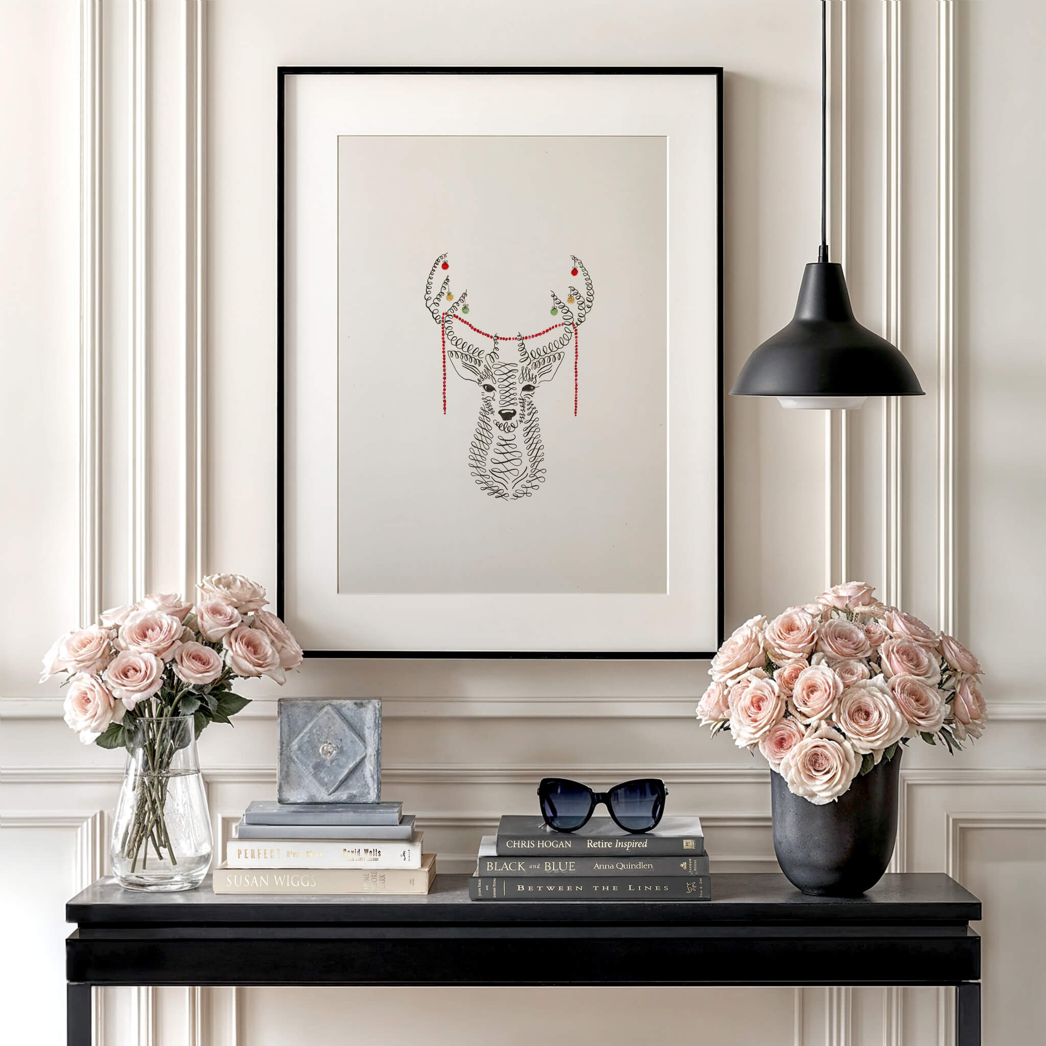 Original pen and ink deer drawing in a black frame with a wide mat above a black console styled with pink roses, stacked books and a black pendant lamp.