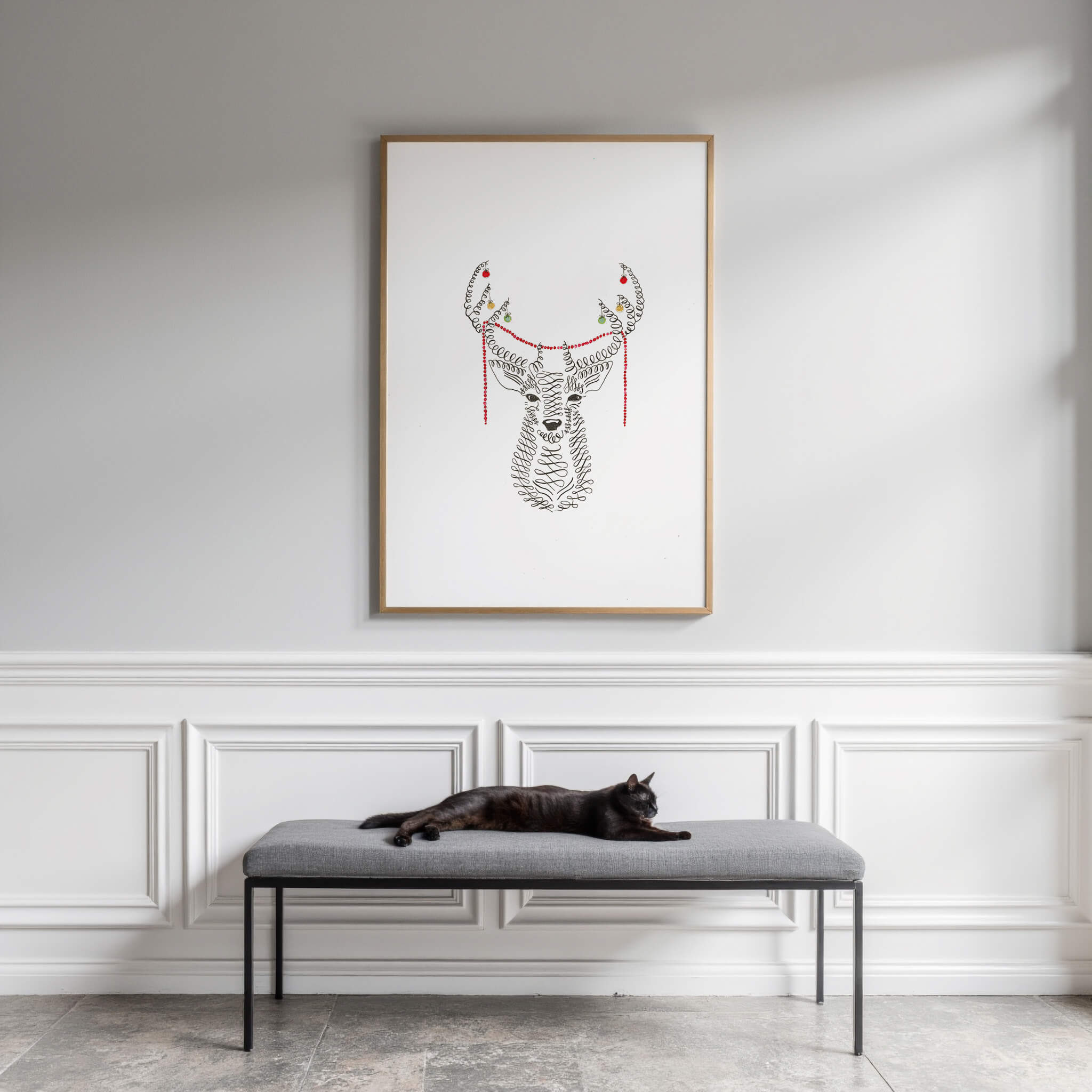 Original calligraphy deer drawing in a thin oak frame above a grey bench where a black cat lounges, against white panelled walls.