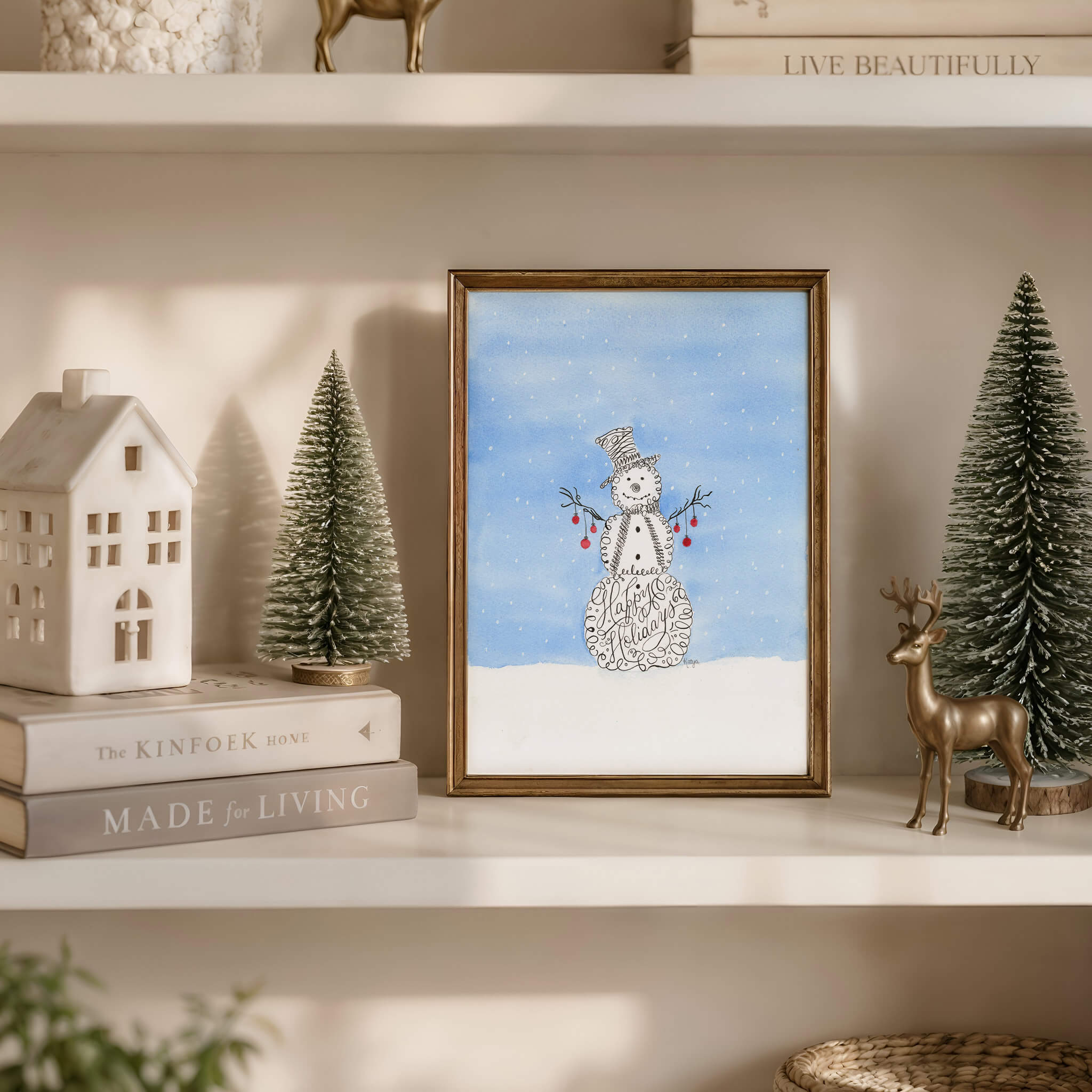 Snowman watercolor painting of a calligraphy snowman in a thin gold frame on a cream shelf with bottle-brush trees, a ceramic house and a brass deer