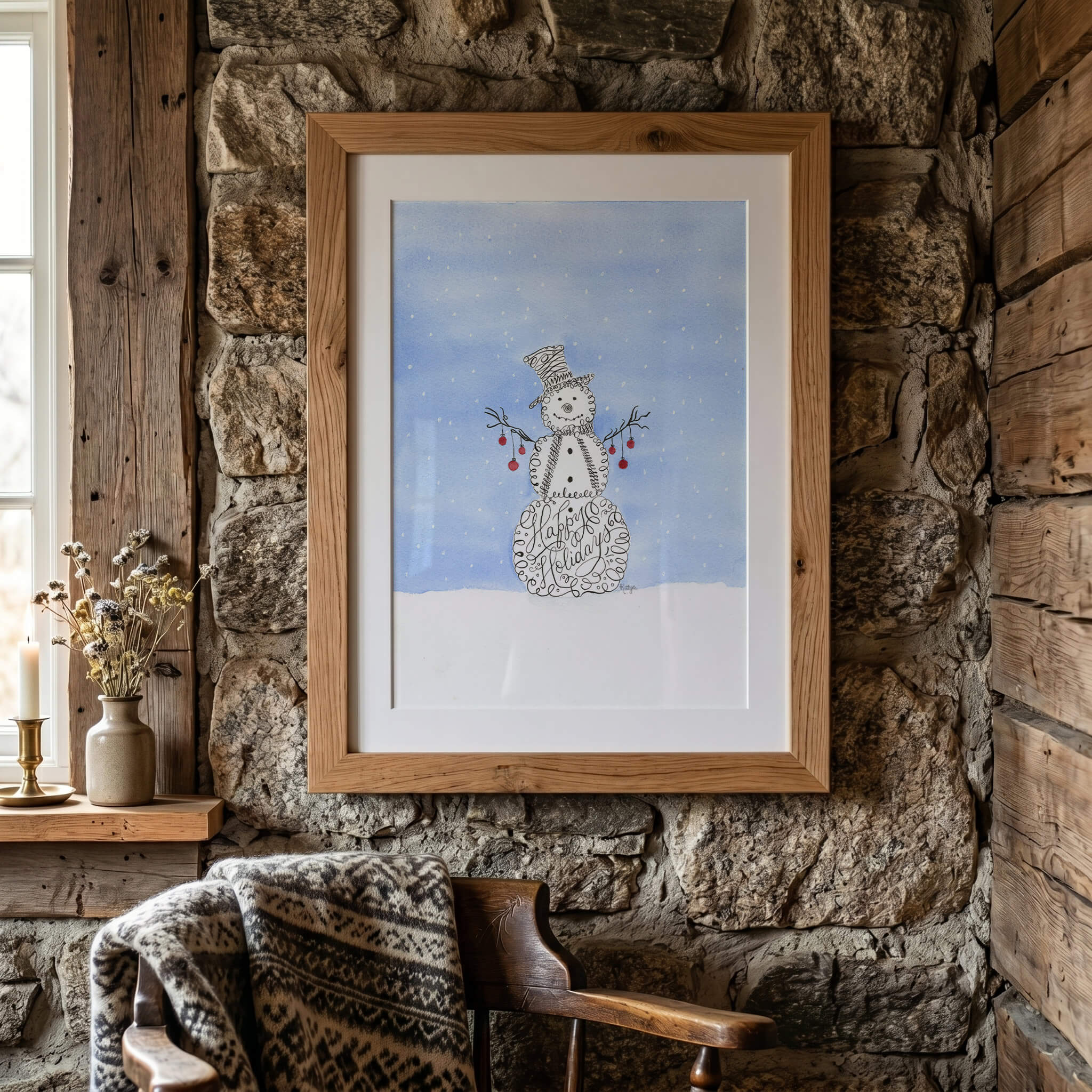 Snowman watercolor painting of a calligraphy snowman in an oak frame with a white mat, hung on a rustic stone wall above a wooden chair draped with a knit blanket