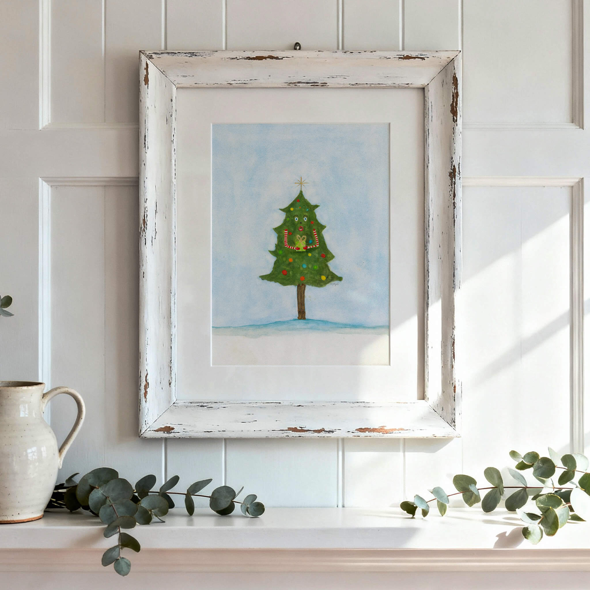 Original Christmas painting of a whimsical watercolor tree holding a gift, in a white distressed frame on a sunny shelf with eucalyptus and a cream pitcher