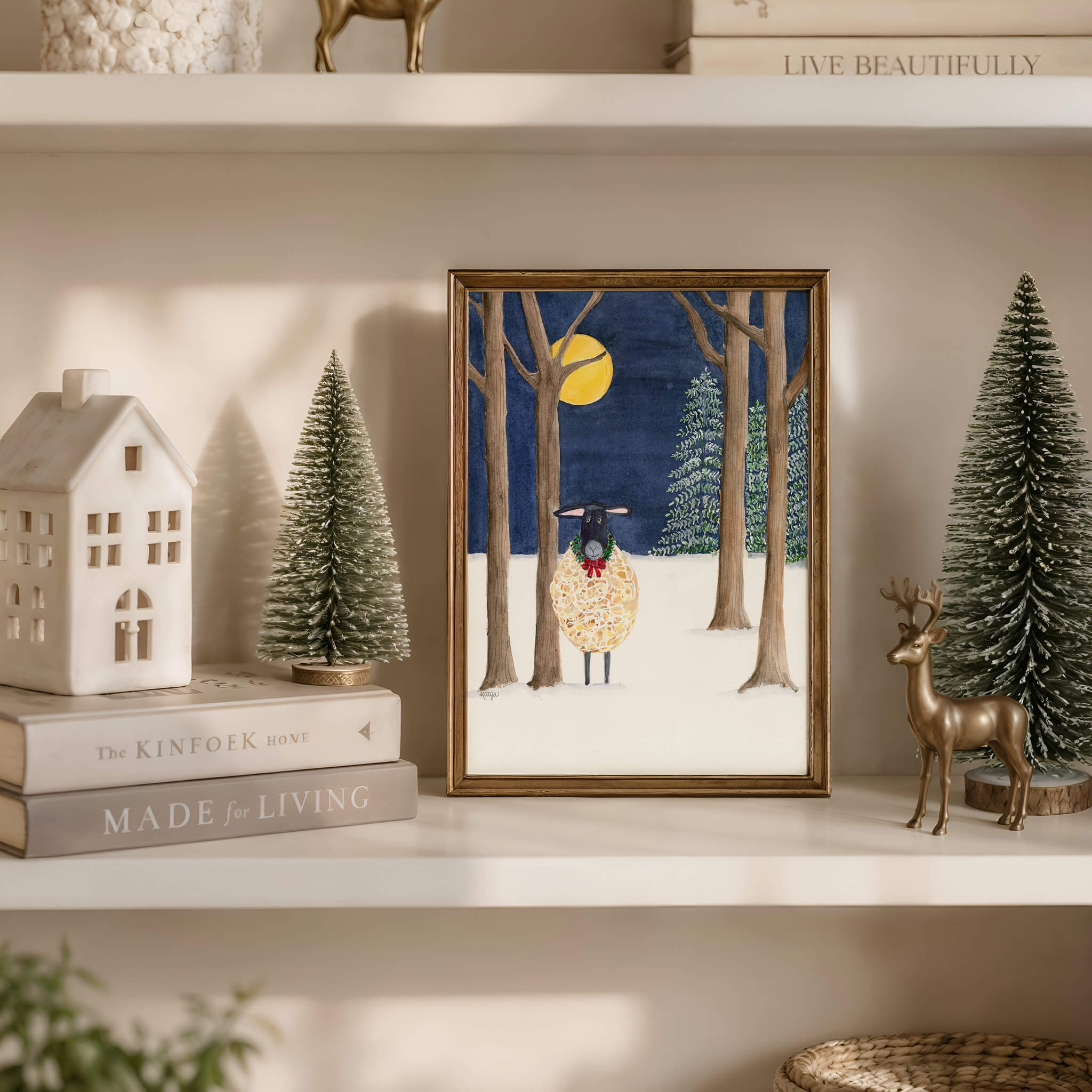 Original sheep painting of a black-faced Suffolk sheep in a green holiday wreath under a full moon, framed on a cream shelf with bottle-brush trees, a ceramic house and a brass deer