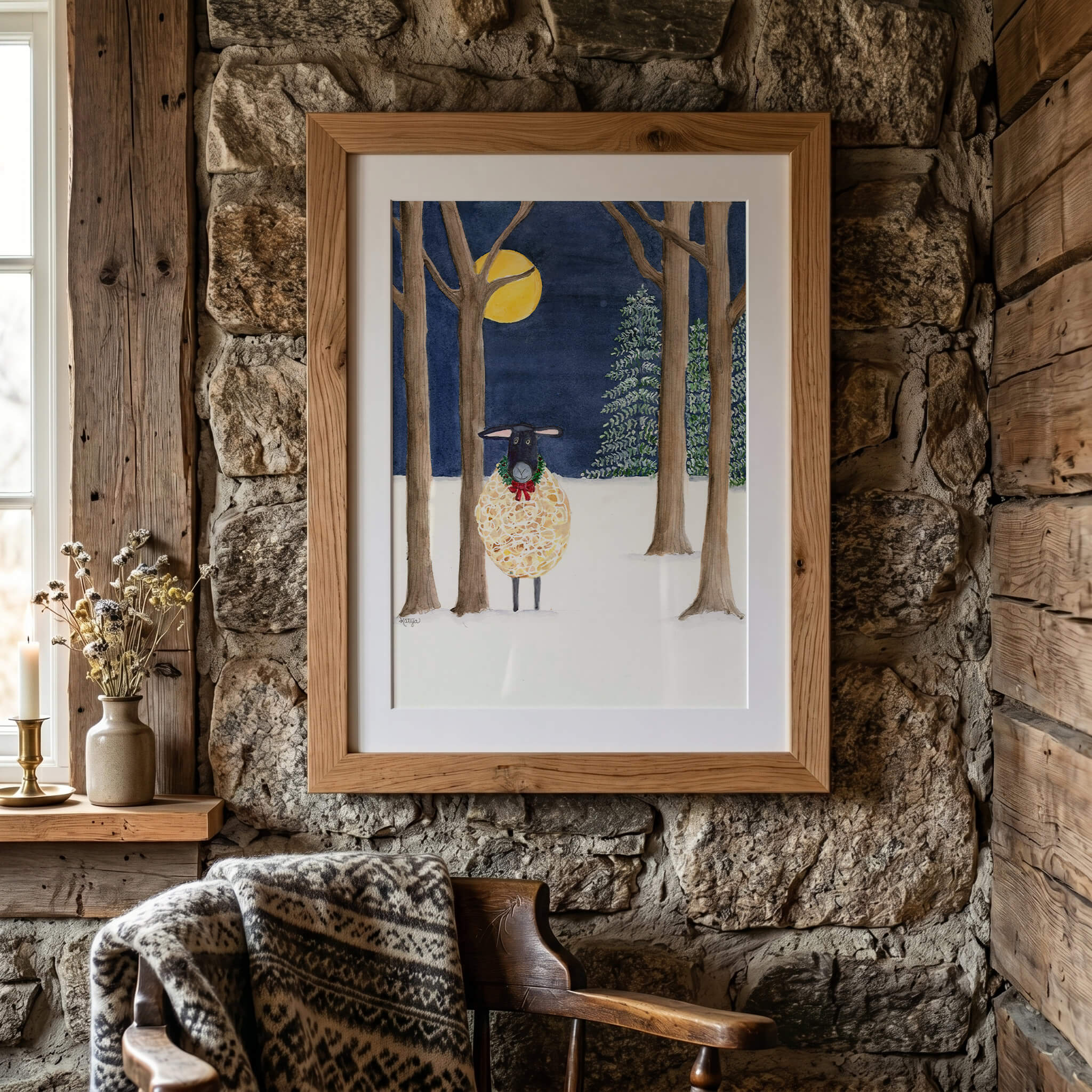 Original sheep painting of a black-faced Suffolk sheep in a green holiday wreath under a full moon, in a matted oak frame on a rustic stone cabin wall above a wooden chair with a knit throw
