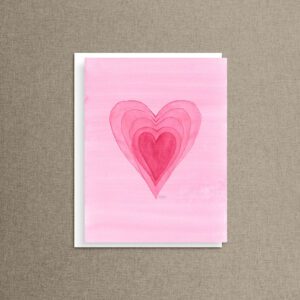 Pink watercolor notecard with nested hearts fading from soft pink to deep rose, propped on a linen background