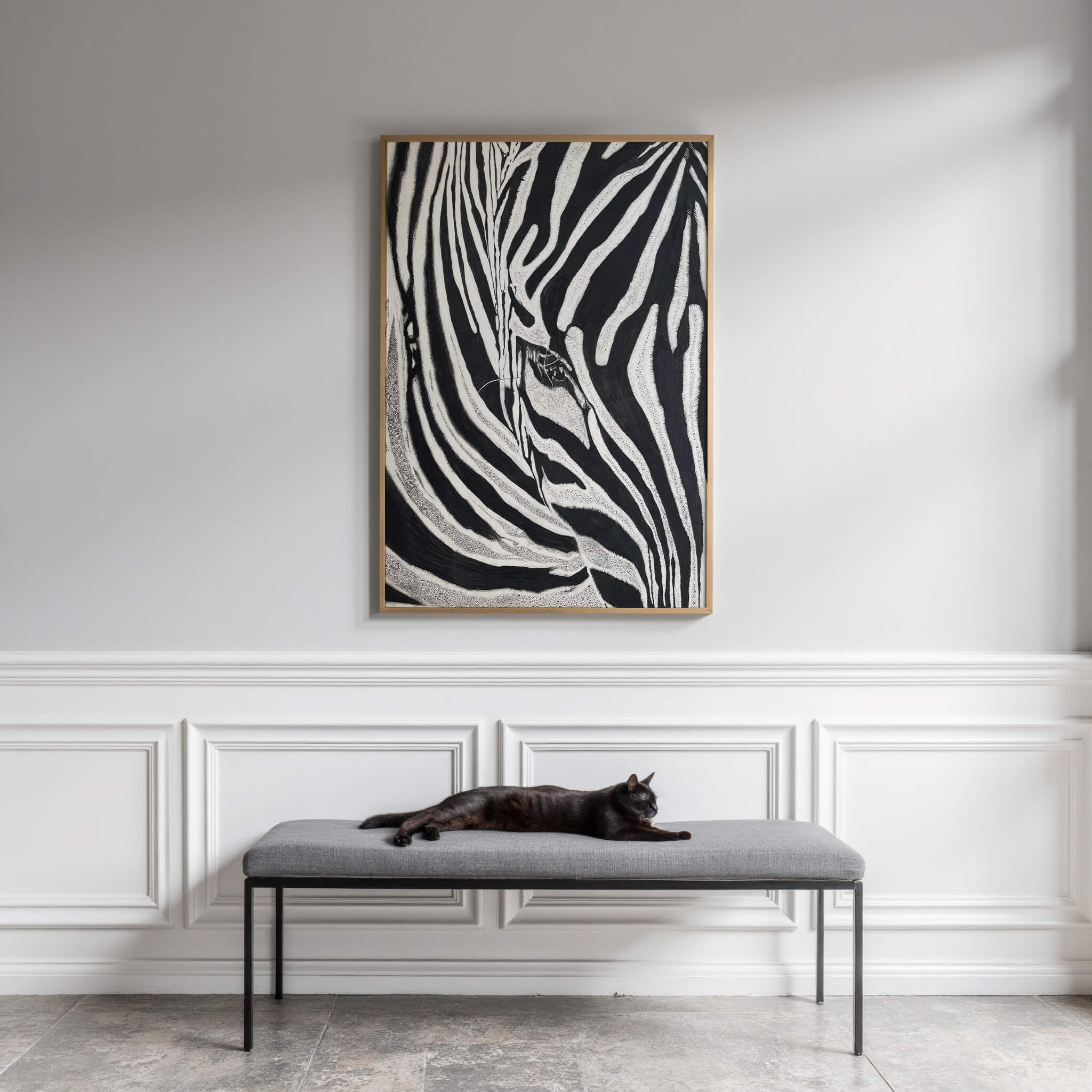 Simply Stripes zebra artwork in an oak frame displayed on a white paneled wall, above a gray bench where a cat rests. The elegant wainscoting, soft natural light, and neutral tones create a refined, gallery-like entryway.