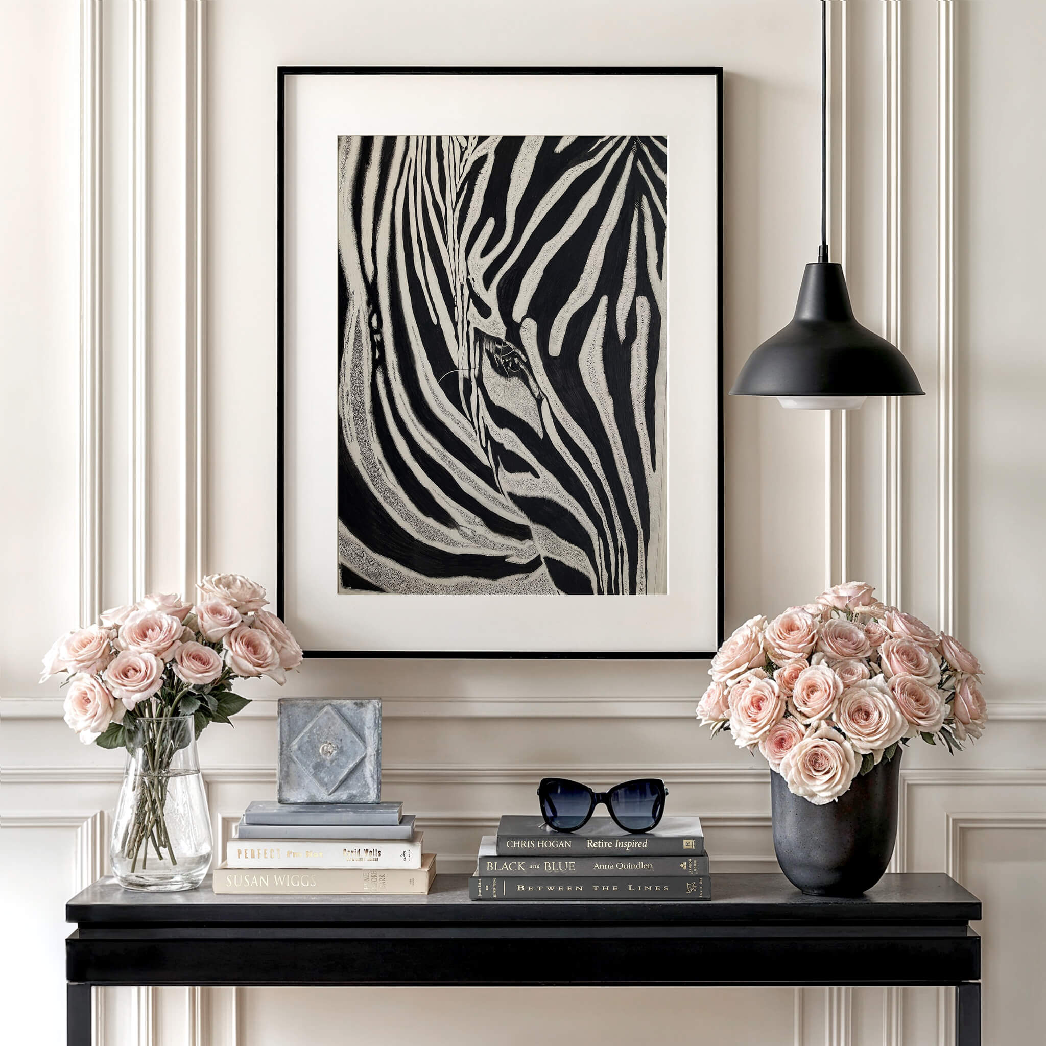 Simply Stripes zebra print in a black frame above a black console table, flanked by pink rose bouquets. Styled with stacked books, sunglasses, a black pendant light, and elegant wall paneling for a chic, feminine touch.