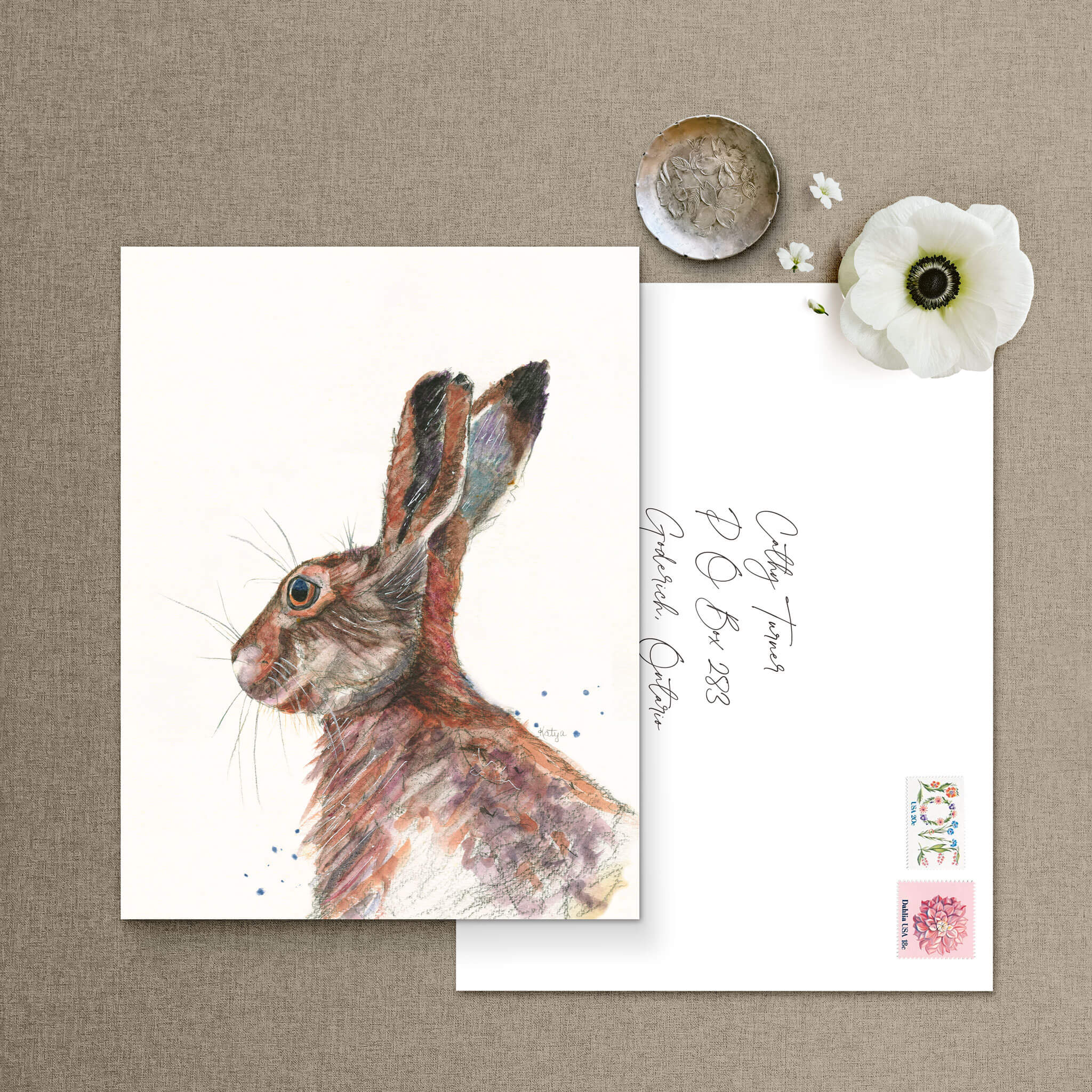 Colorful rabbit note card beside a hand-addressed envelope with vintage flower stamps and a white anemone — watercolor hare art by Cathy Turner.